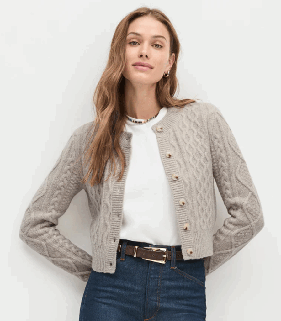 A women wearing a gray cable-knit cardigan over a white shirt, paired with dark wash jeans and a brown belt, delicate beaded necklace and small gold hoop earrings. 