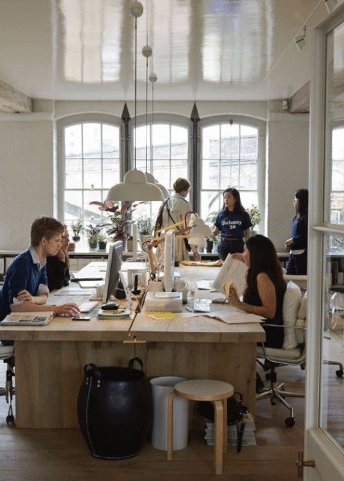 A group of people work at a large wooden desk in a bright office with large windows. Some people are at computers, while others stand and talk.