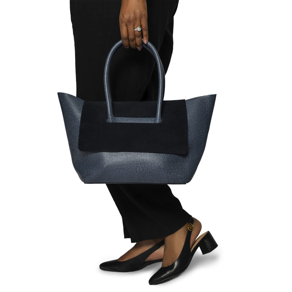 A person holding a blue textured tote bag with a black suede panel and black slingback heels.