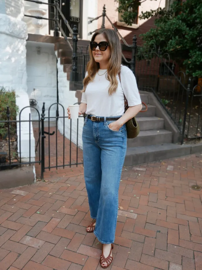 A person wearing a white t-shirt, blue jeans, and brown sandals stands on a brick sidewalk, holding a coffee cup and a bag.