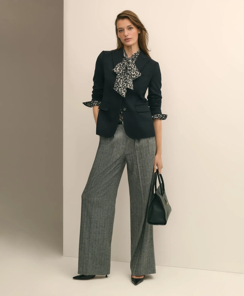 A woman poses in a black blazer, a patterned blouse with a bow, and wide-leg herringbone trousers, holding a black handbag.