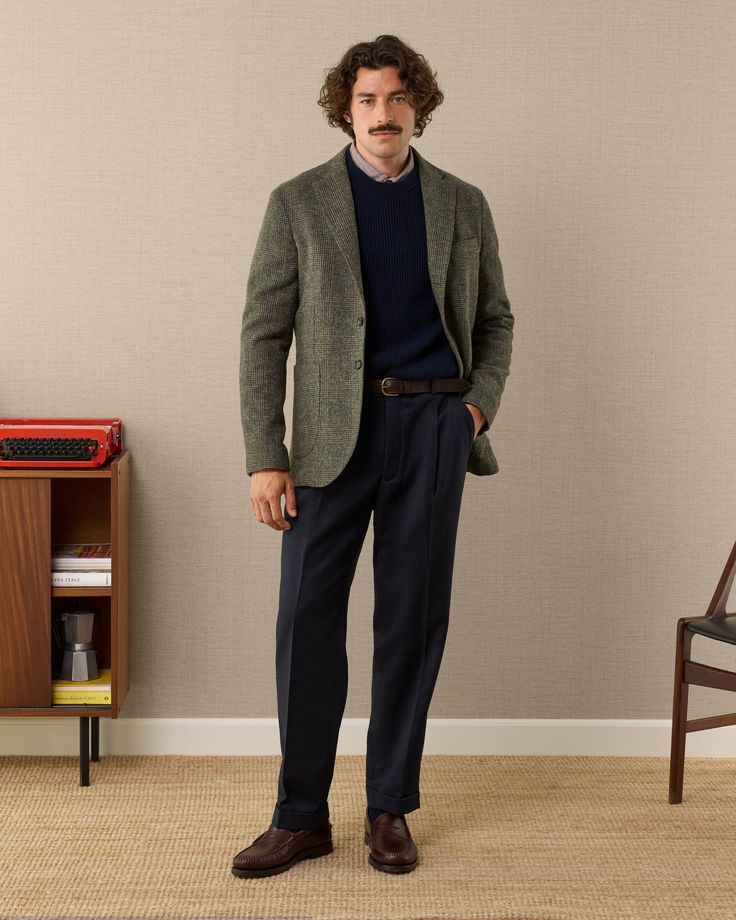 A person wearing a green tweed blazer, a navy cable-knit sweater, a collared shirt, navy trousers, and brown loafers stands against a textured beige wall.