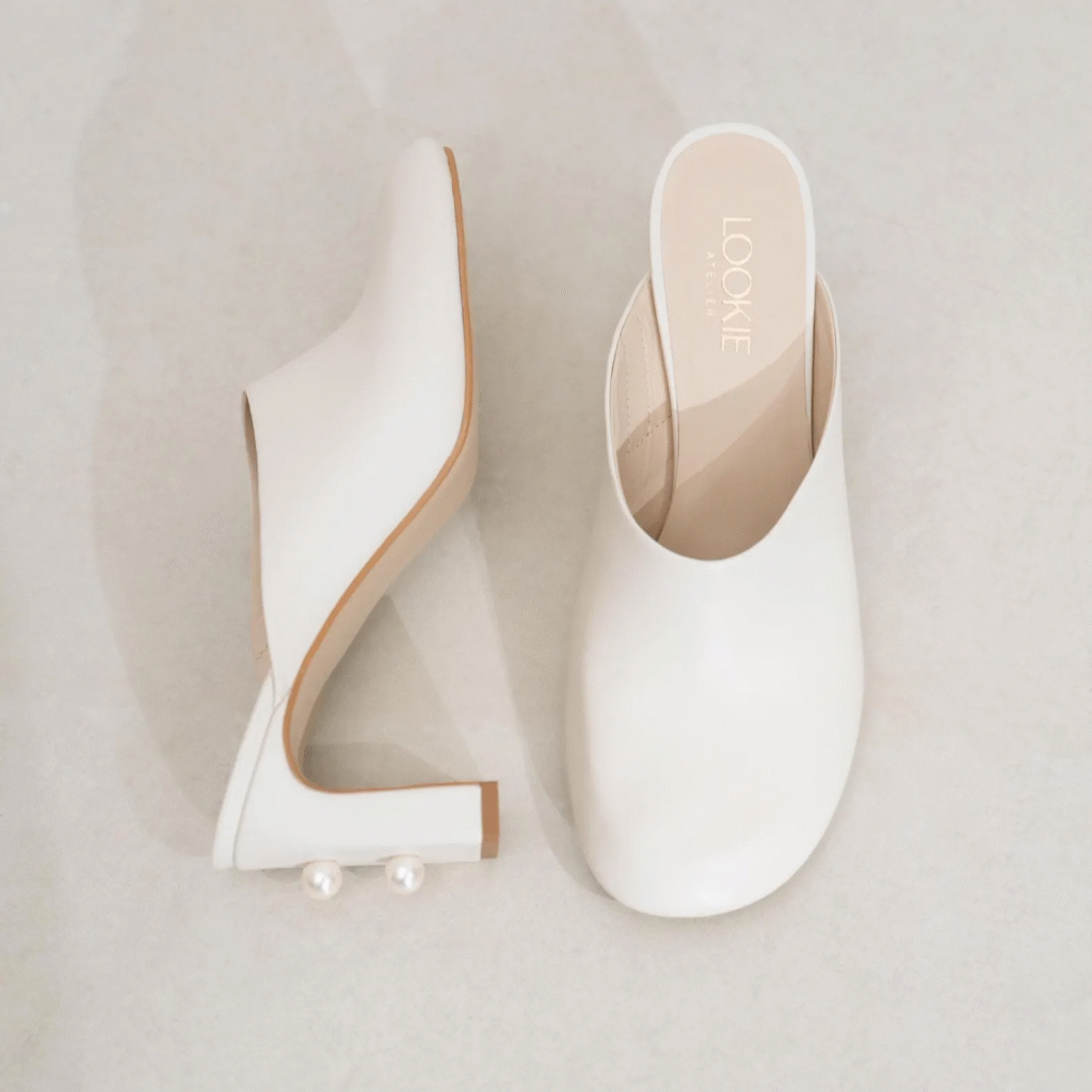 A pair of white leather mules with a square toe and a sculpted heel adorned with two pearl accents.