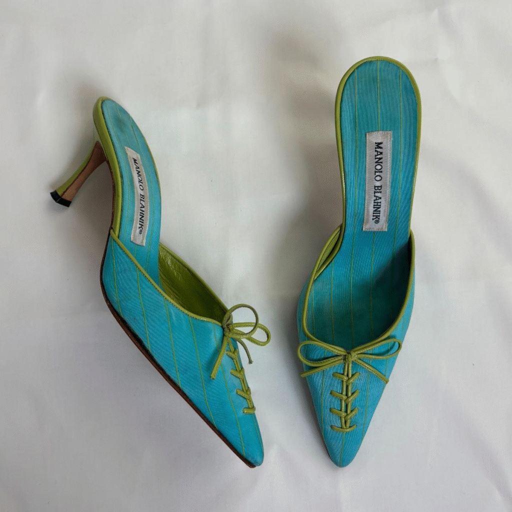 A pair of bright blue Manolo Blahnik mules with lime green trim and laces across the toe. The shoes have pointed toes and stiletto heels.