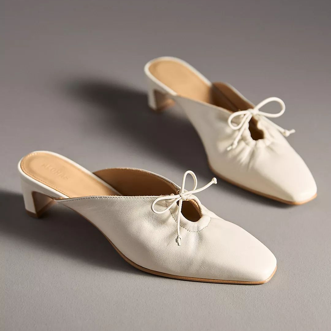 A pair of white leather mules with square toes and delicate white bows on the vamp.