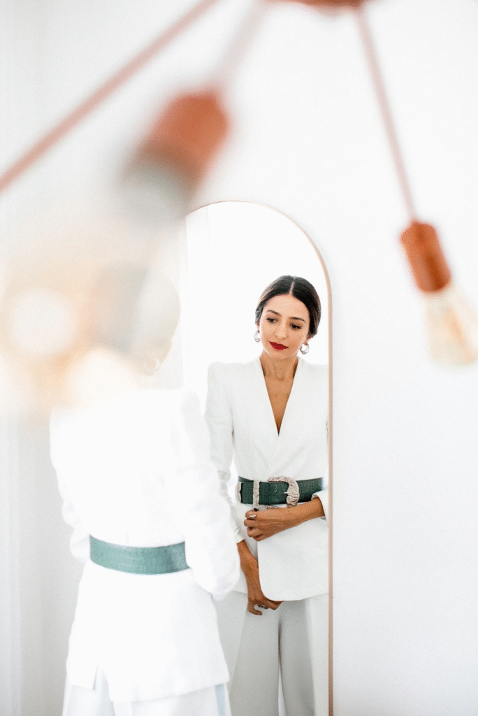 A person in a white blazer and light-colored pants with a wide, dark green belt looks into a mirror.