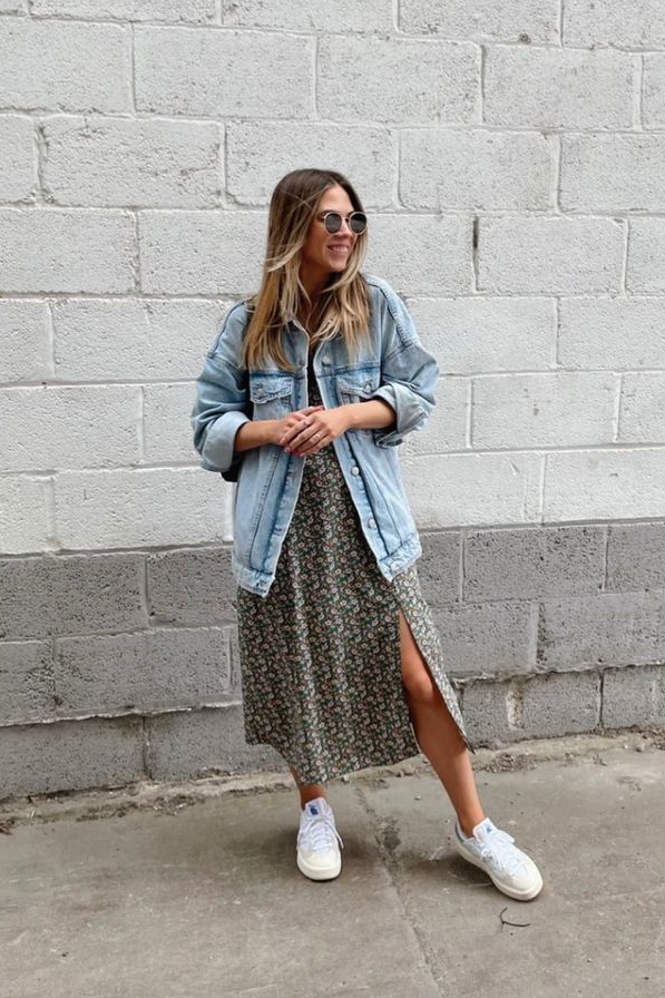 A person wearing a denim jacket over a floral print dress with a thigh-high slit and white sneakers, standing against a white brick wall.