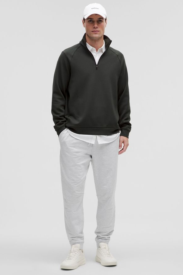 A person wearing a dark green quarter-zip sweatshirt over a white collared shirt, light grey sweatpants, and white sneakers, with a white baseball cap.