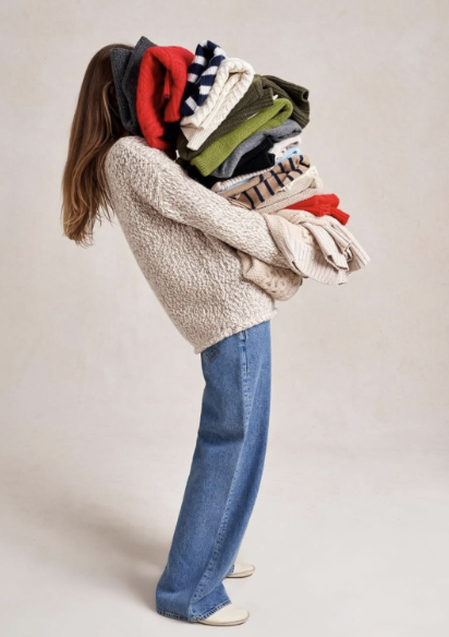 A person wearing a cream-colored sweater and blue jeans is holding a large stack of folded sweaters in various colors and patterns.