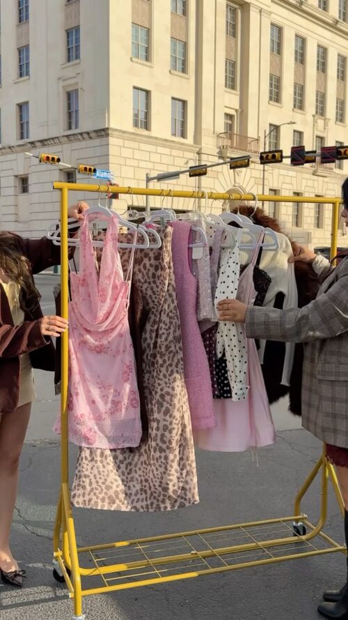 A yellow clothing rack displays various dresses, including a pink floral halter dress and a leopard print dress, on a street in front of a building.