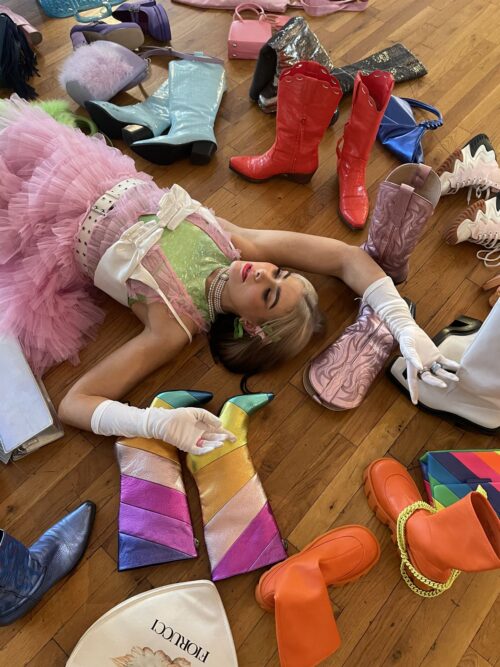 A person in a pink tutu and green and pink top lies on a wooden floor surrounded by various colorful boots, shoes, and handbags.