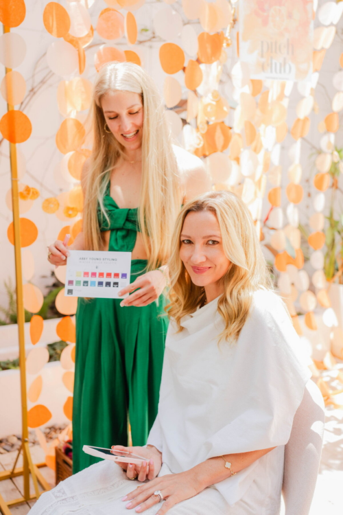 A person in a green outfit holds up a card with swatches of different colors to another person draped in white fabric.