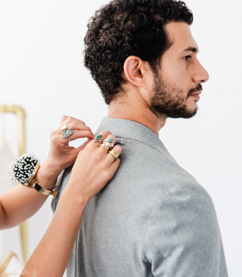 A tailor pins the collar of a gray suit jacket on a man with curly hair.